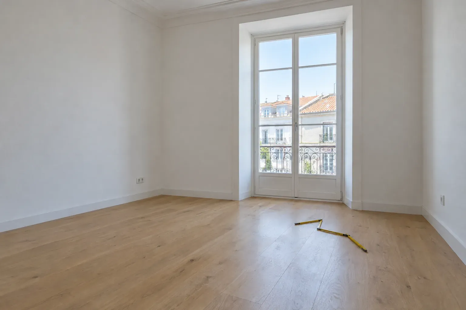Sala vazia de um apartamento, com piso de madeira, uma janela alta e uma fita métrica no chão.